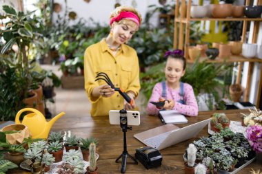 Bir anne ve kız, yeşillik ve aletlerle çevrili bir bitki dükkanında bahçıvanlık dersi çekiyorlar.