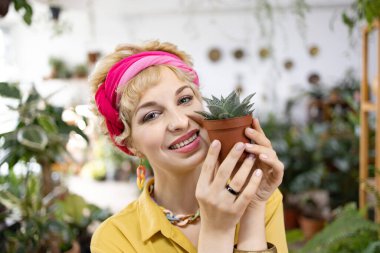 Gülümseyen bir kadın, parlak, sera ortamında küçük bir saksı sulu bitki tutuyor.