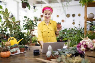 Bir kadın bir fabrikada dizüstü bilgisayarı üzerinde çalışırken küçük, sulu bir saksı tutuyor.