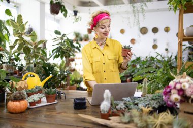 Sarı tişörtlü bir kadın, yeşilliklerle çevrili bir fabrikada dizüstü bilgisayarla çalışırken sulu bir şey tutuyor.