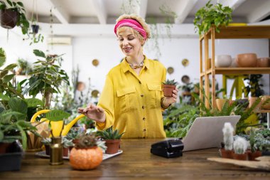 Sarı gömlekli bir kadın, yeşilliklerle çevrili bir bitki dükkanında küçük bir sulu şey tutuyor.