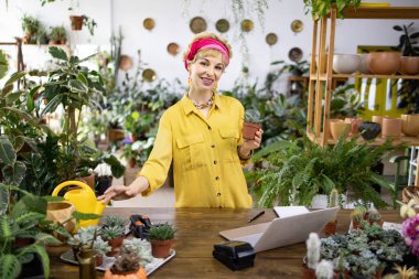 Sarı tişörtlü bir kadın elinde küçük bir saksı suyu tutarken gülümsüyor.