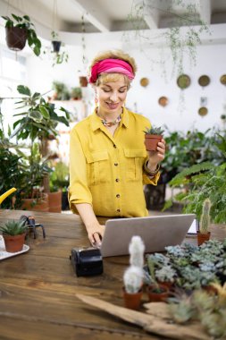 Diz üstü bilgisayarda çalışan bir kadın parlak, davetkar bir bitki dükkanında küçük bir saksı suyu tutuyor.