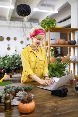 Gülümseyen bir kadın, yeşilliklerle çevrili bir fabrikadaki ahşap tezgahta dizüstü bilgisayarda çalışıyor.