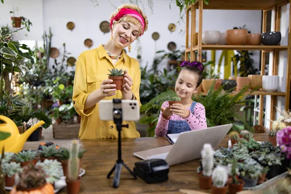 Bir kadın ve bir kız, bir çiçekçide video çekerken ellerinde küçük saksılar tutuyorlar.