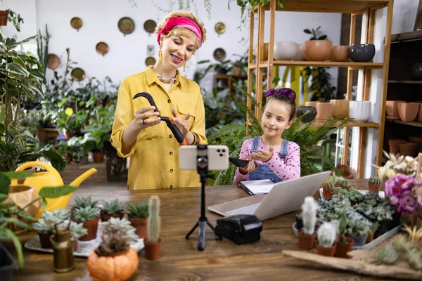 Bir kadın ve bir kız, yeşillik ve aletlerle çevrili bir bitki dükkanında bahçıvanlık videosu çekiyorlar.