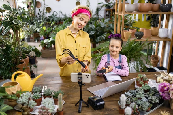 Bir kadın ve bir kız bahçeyle ilgili bir dersi birlikte çekiyorlar. Etrafı bitki ve bahçe aletleriyle çevrili.