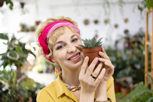 Gülümseyen bir kadın, parlak, sera ortamında küçük bir saksı sulu bitki tutuyor.