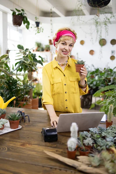 Gülümseyen bir kadın, parlak, davetkar bir bitki dükkanında dizüstü bilgisayar üzerinde çalışırken küçük bir sulu bir şey tutar.