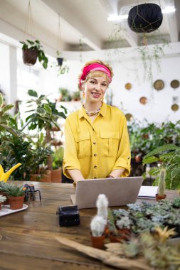 Gülümseyen bir kadın, parlak, davetkar bir dükkanda, yemyeşil çiçeklerle çevrili bir dizüstü bilgisayarda çalışır.