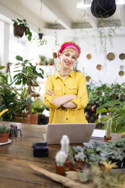 Gülümseyen bir kadın çiçekçisinde kolları çaprazlanmış, yeşilliklerle çevrili duruyor.