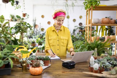 Sarı tişörtlü bir kadın, parlak bir dükkandaki yemyeşil çiçeklerle çevrili bir dizüstü bilgisayarda çalışıyor.