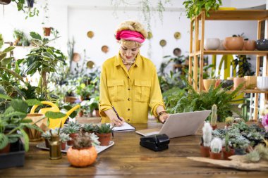 Sarı gömlekli bir kadın, bitkilerle çevrili ahşap bir masada çalışıyor, deftere yazıyor ve dizüstü bilgisayar kullanıyor.