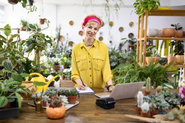 Gülümseyen bir kadın, yemyeşil bitkiler ve sularla çevrili ahşap bir masada çalışıyor.