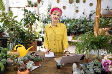 Gülümseyen bir kadın ahşap tezgahta çalışıyor. Etrafı yemyeşil bitkiler ve sularla çevrili.