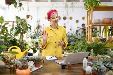 Sarı tişörtlü bir kadın bitkilerle dolu bir dükkanda video görüşmesi yaparken el sallıyor.