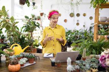 Sarı tişörtlü bir kadın, çiçekçisinden online olarak bahçıvanlık dersi veriyor.