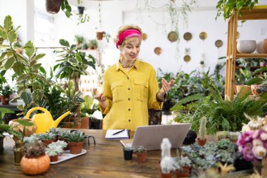 Sarı gömlekli bir kadın, yemyeşil ve dizüstü bilgisayarla çevrili bir bitki dükkanında konuşurken el kol hareketi yapıyor.