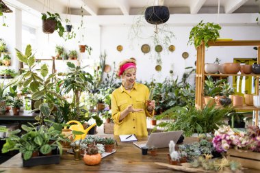 Sarı tişörtlü bir kadın, yeşillik ve dizüstü bilgisayarla çevrili bir fabrikada kameraya kaktüs gösteriyor.