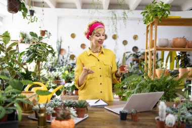 Sarı gömlekli bir kadın kaktüs tutuyor ve parlak, havadar bir odada çiçeklerle ilgili bir ders veriyor.