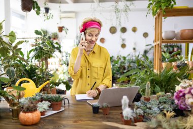 Sarı tişörtlü bir kadın, çevresi yeşilliklerle çevrili bir fabrikada dizüstü bilgisayarla çalışırken telefonla konuşuyor.