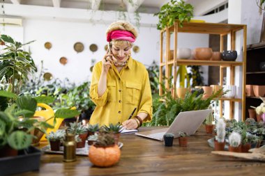 Sarı tişörtlü bir kadın bir fabrikada dizüstü bilgisayar üzerinde çalışırken telefonla konuşuyor.