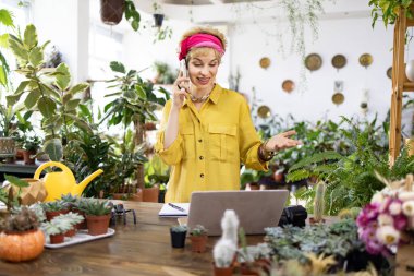 Sarı tişörtlü bir kadın telefonda konuşurken etrafı yemyeşil bitkiler ve sulu bitkilerle çevrili.