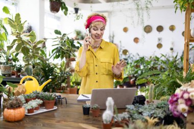 Sarı tişörtlü bir kadın, yemyeşil bir çiçekçide telefonla konuşuyor.