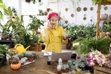 Sarı tişörtlü bir kadın bir çiçekçide telefonda, başparmağını kaldırıyor.