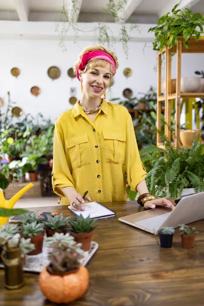 Gülümseyen bir kadın, bitkilerle, sulu sularla ve laptopla çevrili bir masada çalışıyor. Hoş bir ortam yaratıyor.