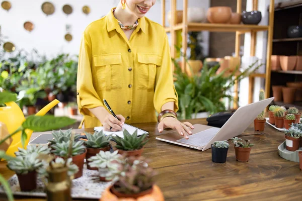 Sarı tişörtlü bir kadın dizüstü bilgisayarda çalışır ve etrafını bitkiler ve sularla çevrili notlar alır.