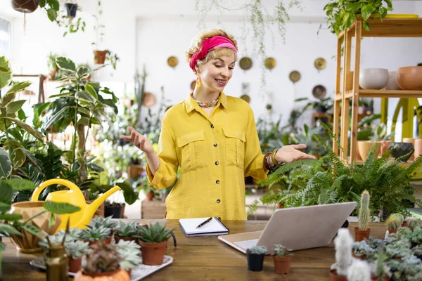 Sarı tişörtlü bir kadın laptopuyla çalışırken etrafı yemyeşil çiçeklerle çevrili.
