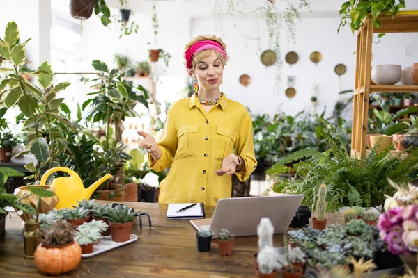 Sarı gömlekli bir kadın, yemyeşil ve sulu çiçeklerle çevrili bir bitki dükkanında dikilirken el kol hareketi yapar.