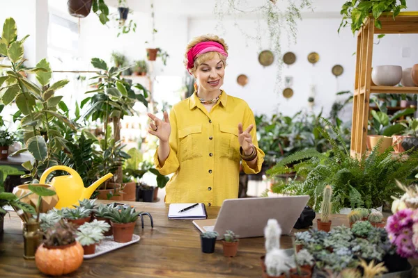 Bir kadın, parlak bir dükkanda bitkilerle çevrili bir dizüstü bilgisayarın önünde konuşurken el kol hareketi yapar.