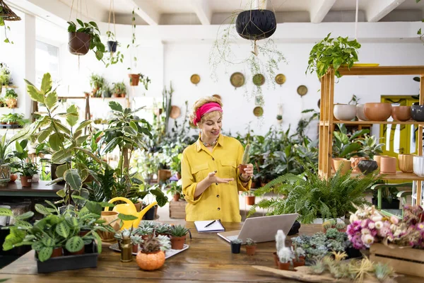 Sarı tişörtlü bir kadın, yeşillik ve dizüstü bilgisayarla çevrili bir fabrikada kameraya kaktüs gösteriyor.