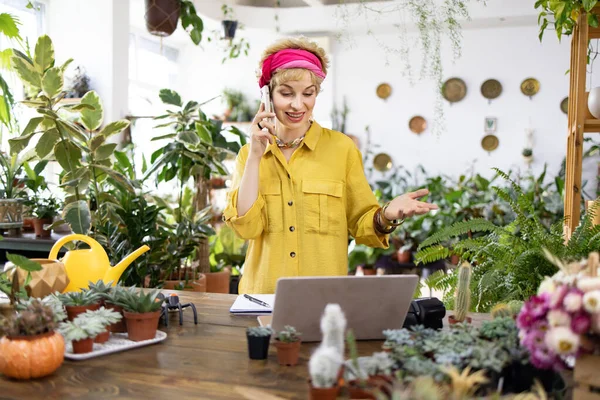 Sarı tişörtlü bir kadın telefonda konuşurken etrafı yemyeşil bitkiler ve sulu bitkilerle çevrili.