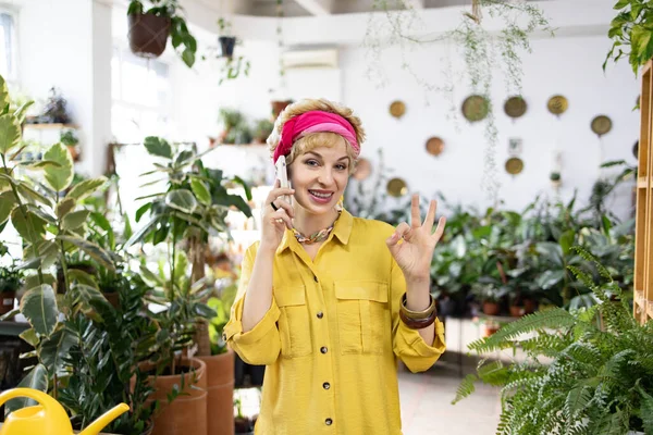 Sarı tişörtlü bir kadın telefonda konuşurken gülümsüyor ve çiçekçide 