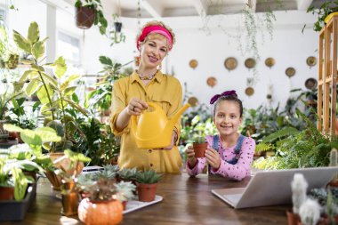 Bir kadın ve bir kız, elinde bir su kovası ve bitki dolu bir odada küçük bir kaktüs tutarken gülümsüyorlar.