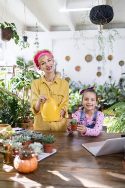 Bir kadın ve bir kız, elinde bir su kovası ve bir bitki dükkanında küçük bir kaktüs tutarken gülümsüyorlar.