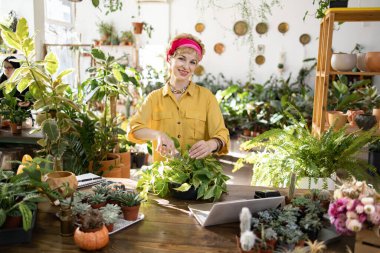 Bir kadın çiçekleri budarken gülümsüyor. Parlak, bitki dolu bir serada.