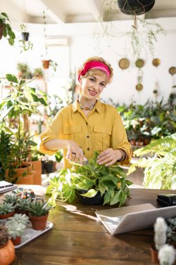 Gülümseyen bir kadın, yemyeşil çimlerle çevrili, parlak, havadar bir serada çiçek budar.