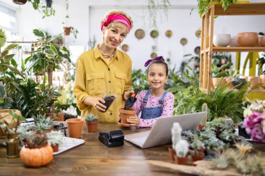 Bir kadın ve genç bir kız, yeşillik dolu, rahat, aydınlık bir dükkanda saksı bitkilerinin tadını çıkarıyorlar.