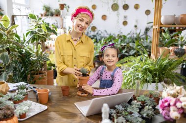 Bitkilerle uğraşırken bir kadın ve genç bir kız gülümsüyor, aile ve doğa için iç açıcı bir sahne yaratıyor.