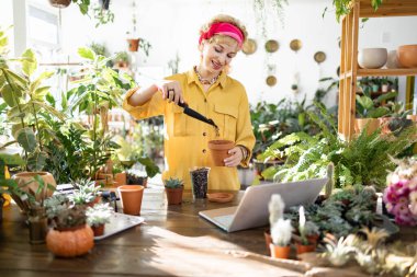 A woman smiles while repotting a plant, surrounded by lush greenery and using a laptop for guidance
