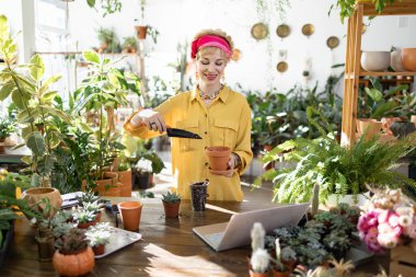 Sarı tişörtlü ve pembe başörtülü bir kadın, yemyeşil bir çömleğin içine sulu suşlar ekiyor.