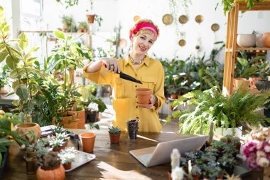 A smiling woman is planting a flower in a pot, surrounded by various plants in a bright, sunny room
