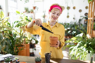 Gülümseyen bir kadın, parlak, güneşli bir odada çeşitli bitkilerle çevrili bir saksıya çiçek dikiyor.