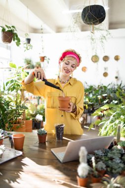 A woman in a yellow shirt and pink headscarf is planting succulents in a terracotta pot, surrounded by lush greenery