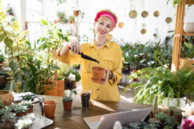 Gülümseyen bir kadın, parlak, güneşli bir seraya sulu bir bitki dikerken görülüyor.