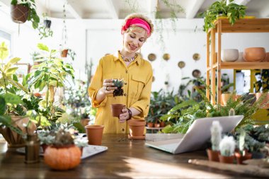 A woman smiles while working on her plants, surrounded by greenery and using a laptop